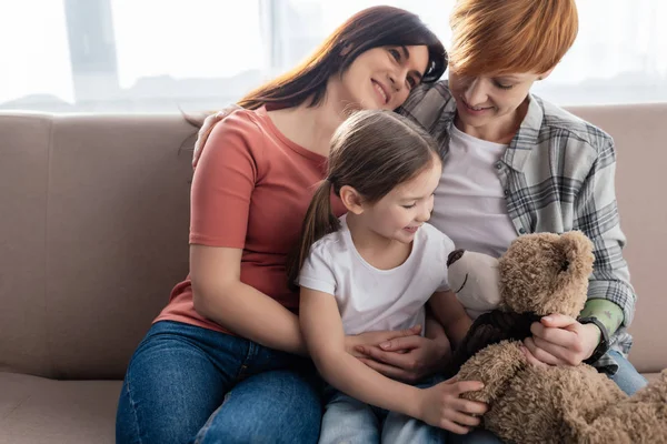 Улыбающиеся Однополые Родители Сидят Рядом Дочерью Плюшевым Мишкой Диване — стоковое фото