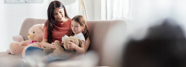 Selective focus of kid playing with teddy bear near smiling mother on sofa in living room, panoramic shot 