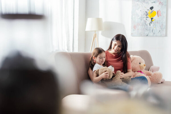 Selective focus of child playing with teddy bear near happy mother on sofa in living room