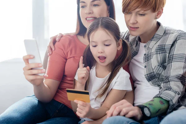 Smiling Same Sex Parents Smartphone Daughter Credit Card Couch Home — Stock Photo, Image