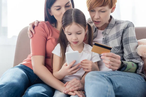 Família Mesmo Sexo Abraçando Filha Usar Smartphone Cartão Crédito Sofá — Fotografia de Stock