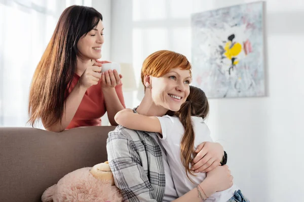 Leende Kvinna Omfamnar Dotter Soffa Nära Förälder Med Kaffekopp Vardagsrummet — Stockfoto