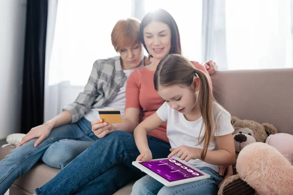 Ekranda elektronik sağlık uygulaması olan dijital tablet kullanan bir çocuk kredi kartı ve yumuşak oyuncakları olan annelerin yanında. 