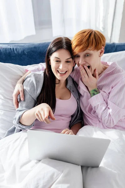 Smiling woman pointing with finger on laptop to shocked girlfriend on bed