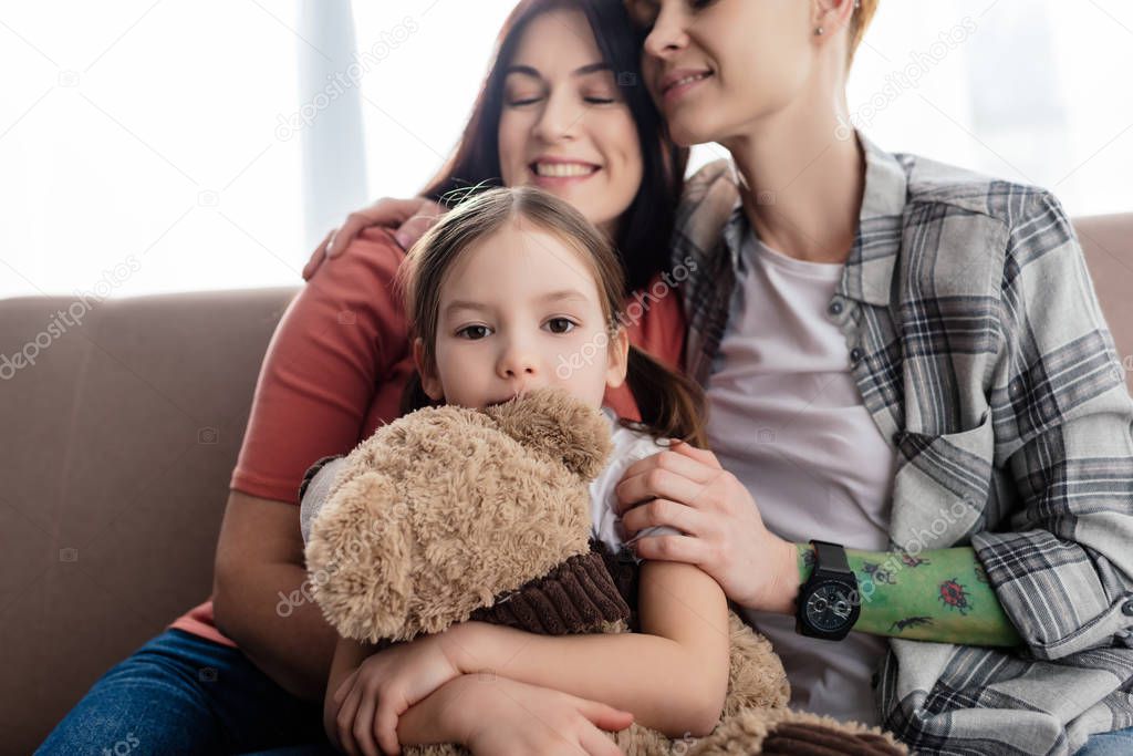 Child with teddy bear looking at camera near smiling same sex parents on couch 