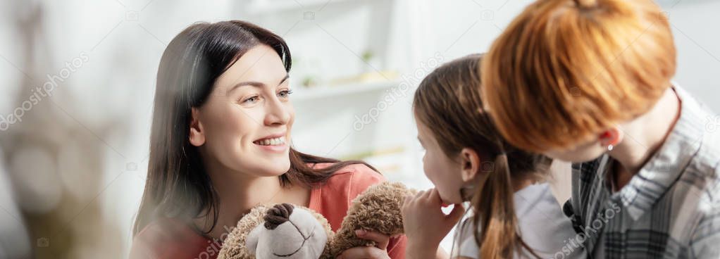 Selective focus of smiling mother holding teddy bear near daughter and parent in living room, panoramic shot