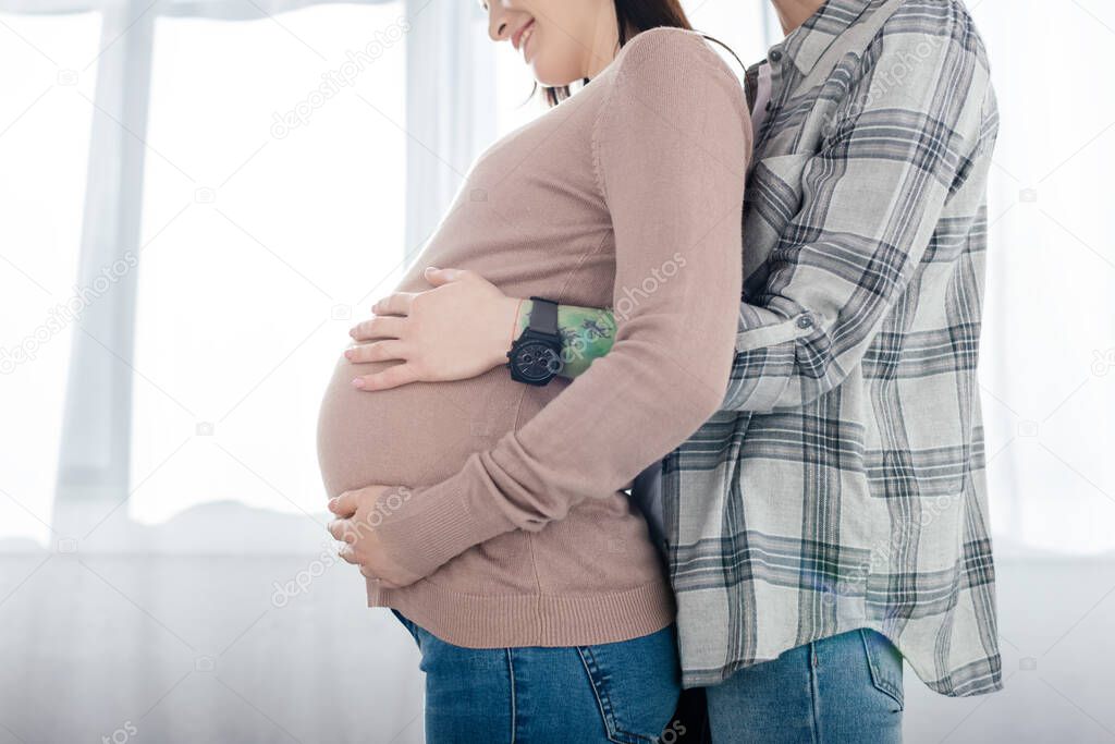 Cropped view of woman touching belly of smiling pregnant girlfriend at home 