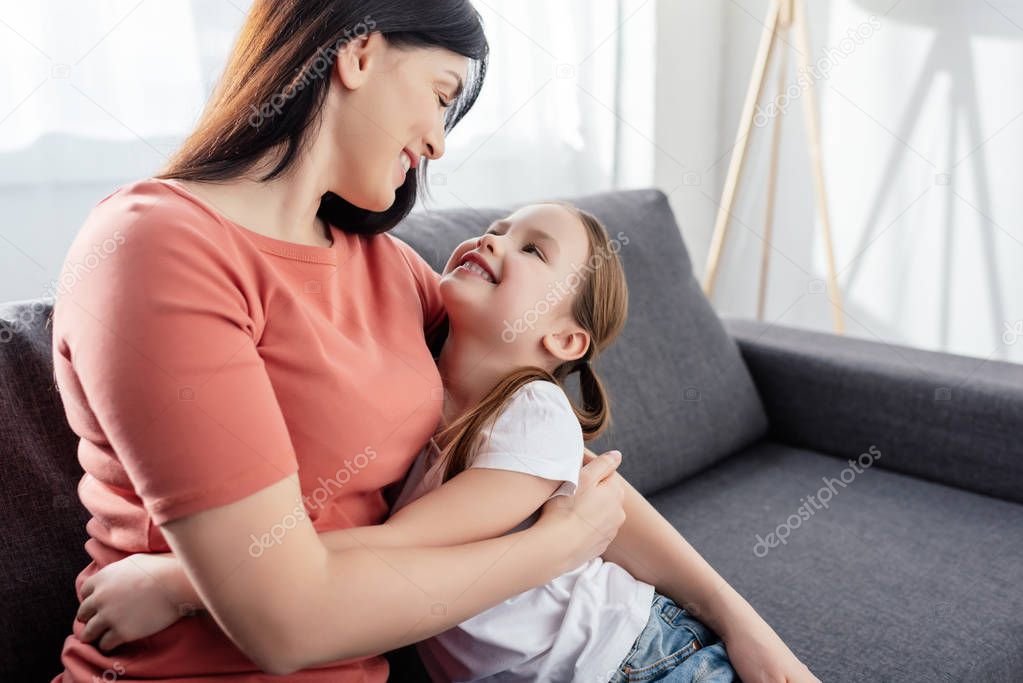 Smiling kid embracing and looking at mother while sitting on couch in living room