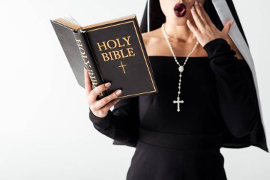 cropped view of shocked nun in black dress reading bible isolated on grey clipart