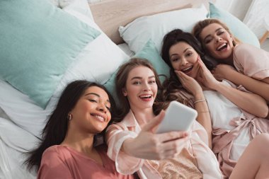 Bekarlığa veda partisinde yatakta dinlenirken selfie çeken heyecanlı, çok kültürlü kadınların yüksek açılı görüntüsü.