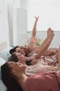 Çok kültürlü kadınların selfie çekip bekarlığa veda partisinde yatakta uzanmaları.