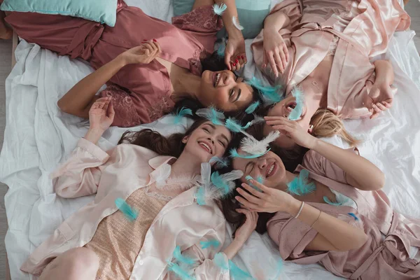 幸せな多民族女性のトップビュー笑顔とベッドの上に寝そべって羽でパーティー — ストック写真