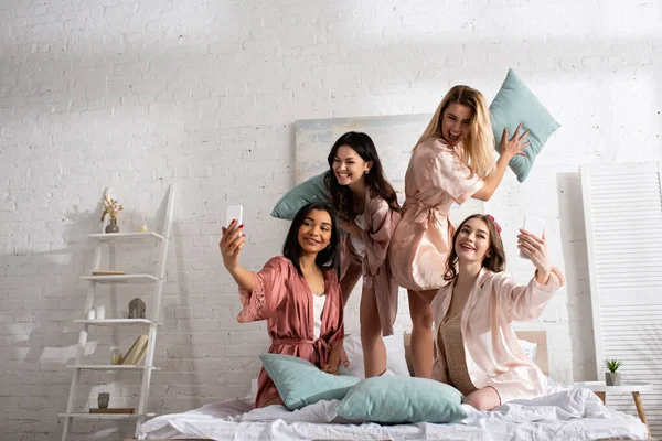 Happy Multicultural Women Smiling Holding Pillows Taking Selfie Smartphones Bed — Stock Photo, Image