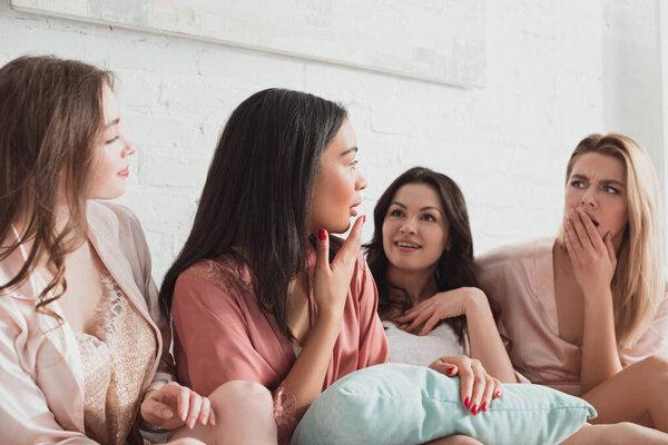 Multicultural women talking and looking at each other with pillow on bed at bachelorette party