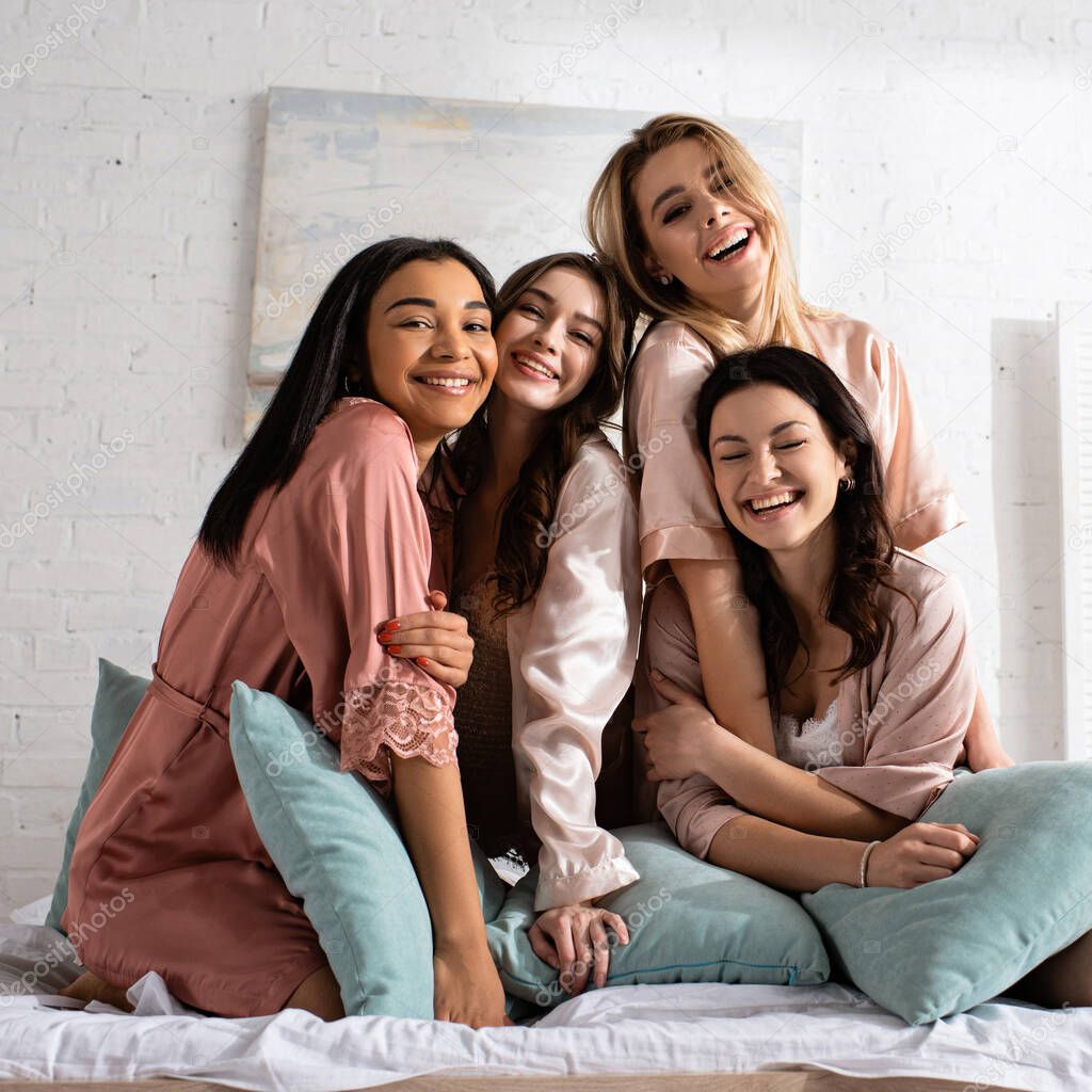 Happy multiethnic friends looking at camera, hugging and smiling together with pillows on bed at bachelorette party