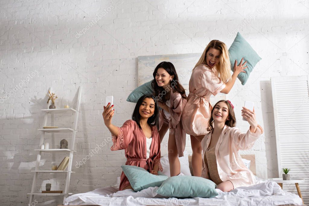 Happy multicultural women smiling, holding pillows and taking selfie with smartphones on bed at bachelorette party