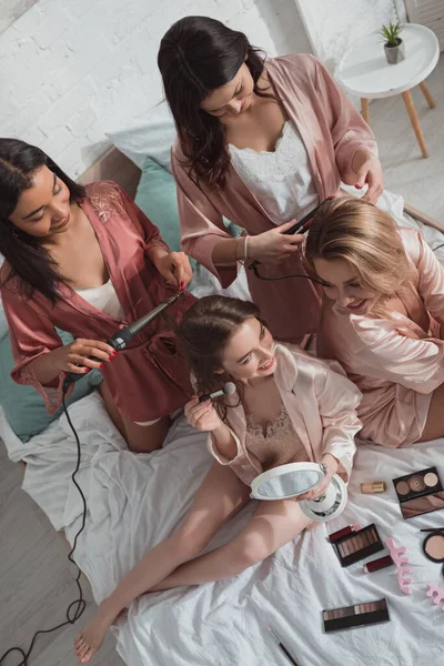 High angle view of multiethnic women together using cosmetics and curling irons on bed at bachelorette party