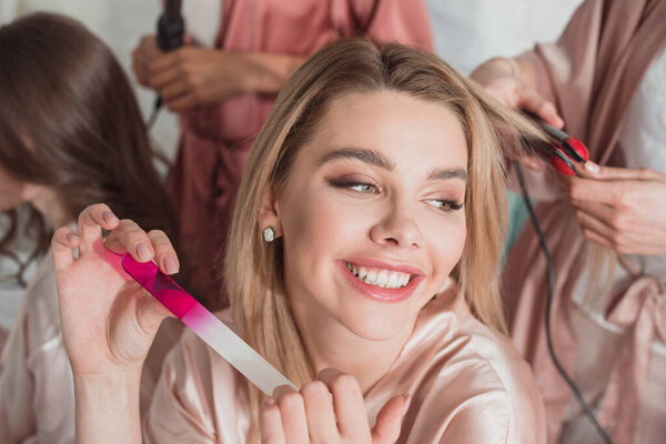 Selective focus of blonde girl holding nail file and smiling with multicultural friends at bachelorette party