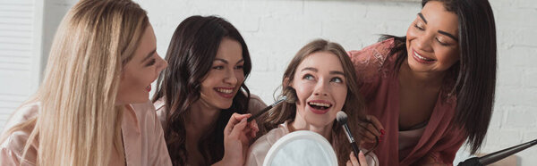 Multicultural women with putting makeup with cosmetic brushes on bride, smiling together in room, panoramic shot