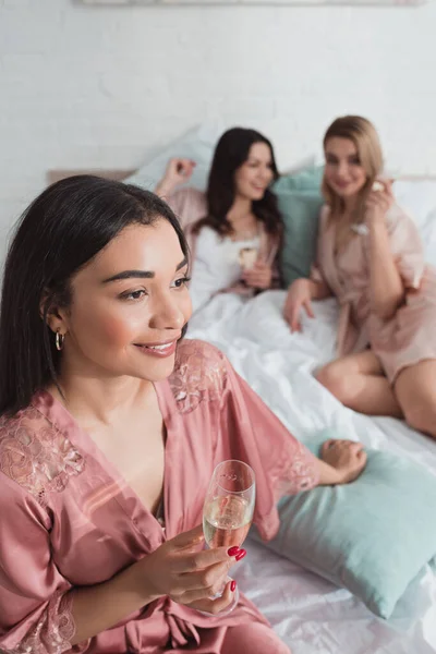 在单身派对上 非洲裔美国女孩带着香槟酒和朋友在床上微笑的选择性焦点 — 图库照片