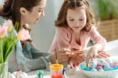 Mutlu anne ve neşeli çocuk çiçeklerin yanında paskalya yumurtası boyuyor.