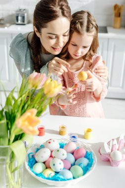 Mutlu anne ve tatlı kızın seçici odağı lalelerin yanındaki paskalya yumurtasına bakıyor. 