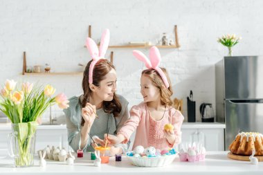 Tavuk yumurtası, dekoratif tavşanlar, paskalya ekmeği ve lalelerin yanında tavşan kulaklı mutlu çocuk ve anne. 
