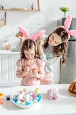 Mutlu bir annenin tavşan kulaklarına dokunup tavuk yumurtası boyaması üzerine seçici bir odaklanma. 