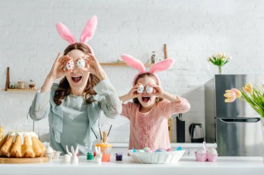 excited mother and daughter in bunny ears covering eyes with painted chicken eggs near tulips and easter bread  clipart