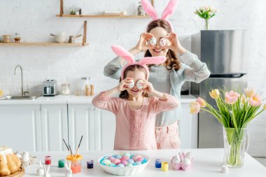 smiling mother and daughter in bunny ears covering eyes with easter eggs near tulips  clipart