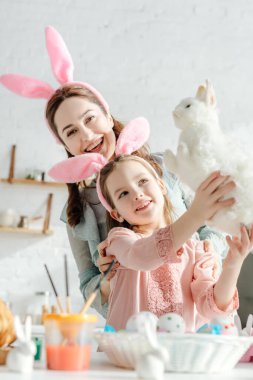 Mutlu anne, tavşan kulaklı çocuğun ellerinde oyuncak tavşana bakıyor. 