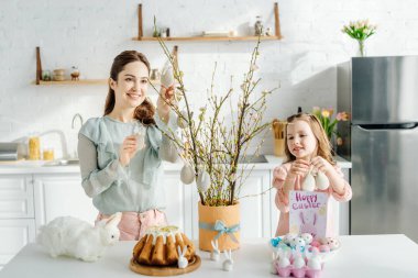 Anne söğüt ve dekoratif tavşanların yanında süslü paskalya yumurtaları tutan sevimli çocuk. 