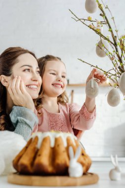 Süslü paskalya yumurtalarıyla söğüt dallarına bakan mutlu anne ve kızın seçici odağı 