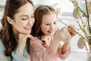 Annesinin yanındaki söğüt dalında süslemeli paskalya yumurtasına dokunan mutlu çocuğun seçici odağı. 