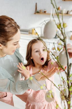 Şok olmuş çocuğun seçici odağı mutlu anne yakınlarındaki söğüt dallarındaki dekoratif paskalya yumurtalarına bakıyor. 