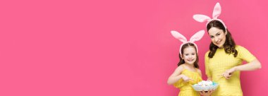 Mutlu anne ve kızın tavşan kulaklarıyla çekilen panoramik görüntüsü pembe üzerinde izole edilmiş paskalya yumurtalarını işaret ediyor. 