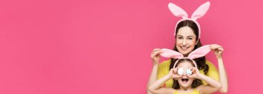 panoramic shot of happy mother touching bunny ears of daughter covering eyes with easter eggs isolated on pink  clipart