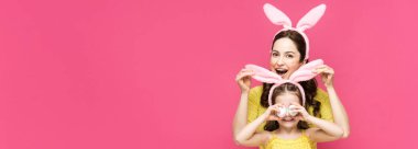 Heyecanlı annenin tavşan kulaklarına dokunuşunun panoramik görüntüsü. Kızının gözleri, pembe üzerine izole edilmiş paskalya yumurtalarıyla kaplanmış. 