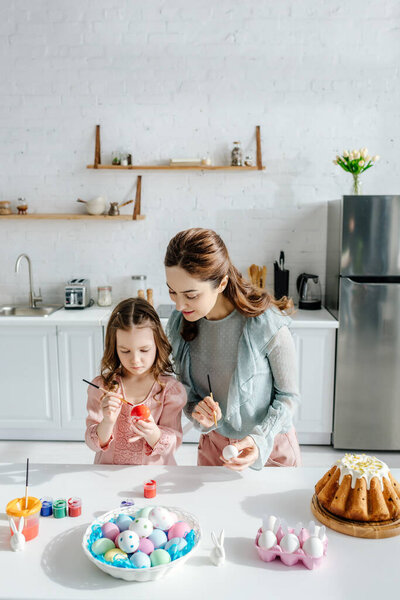 cute child and happy mother painting easter eggs near decorative rabbits and easter bread 