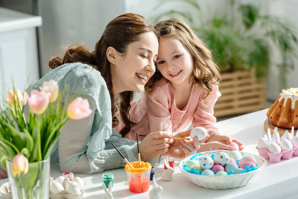 adorable child and happy mother near chicken eggs, decorative rabbits, easter bread and tulips 