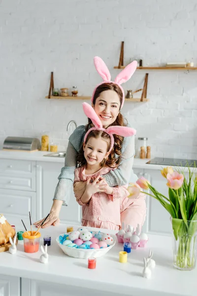 快乐的妈妈把女儿抱在复活节彩蛋旁边的兔子耳朵里 — 图库照片