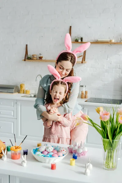 快乐的妈妈把兴奋的女儿抱在复活节彩蛋旁边的小兔子耳朵里 — 图库照片