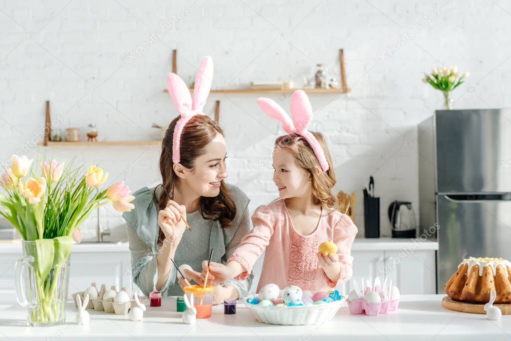 happy child and mother with bunny ears near chicken eggs, decorative rabbits, easter bread and tulips 