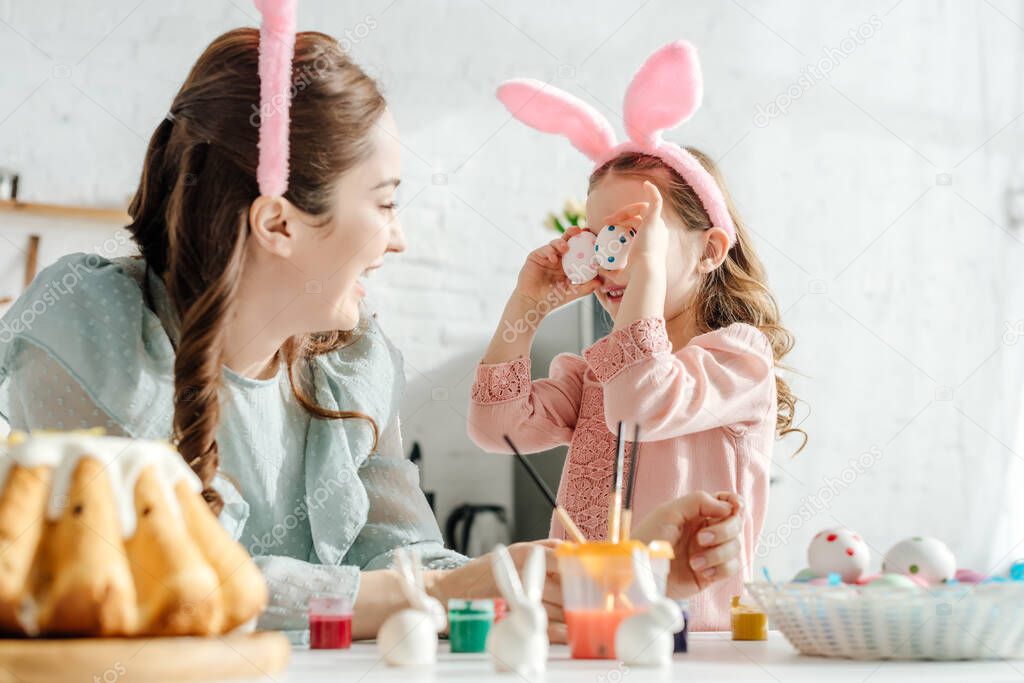 selective focus of happy mother looking at daughter in bunny ears covering eyes with easter eggs 