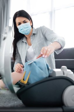 Tıbbi maskeli, elektrik süpürgesinin yanında toz torbası tutan alerjik bir kadın.