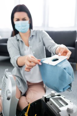 Tıbbi maskeli, elektrik süpürgesinin yanında toz torbası tutan alerjik bir kadın. 