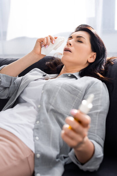 selective focus of allergic woman holding napkin and nasal spray while lying on sofa 