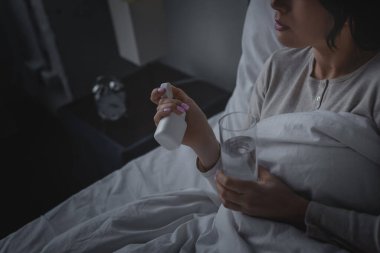Uyku bozukluğu olan bir kadın yatak odasında bir bardak su ve uyku hapıyla bir şişe tutuyor. 