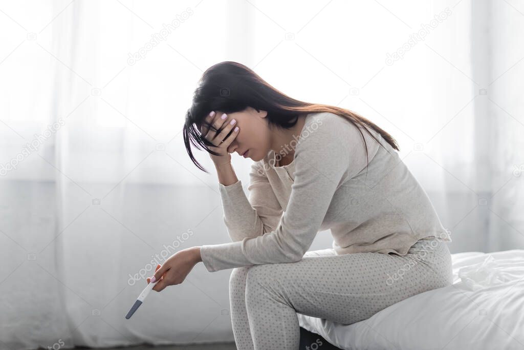 sad woman sitting on bed and holding pregnancy test with negative result 