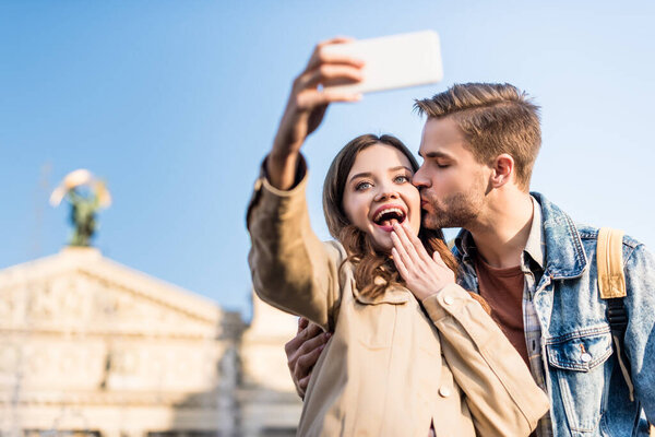 Selective focus of man kissing excited girlfriend taking selfie with smartphone 